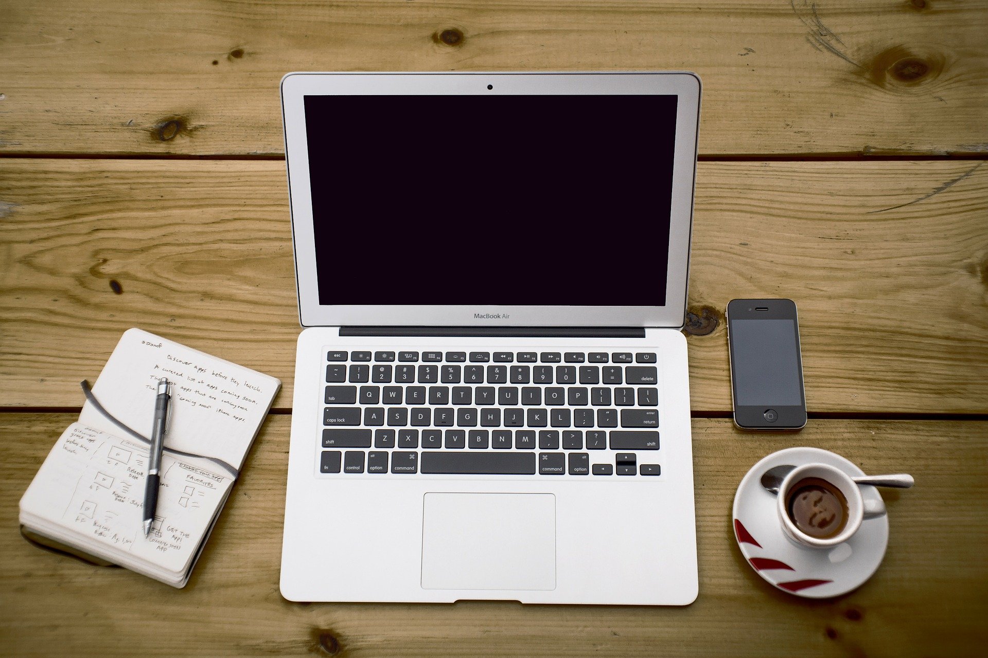 EIn Laptop auf einem Holztisch, daneben notebook, Telefon und Kaffeetasse.