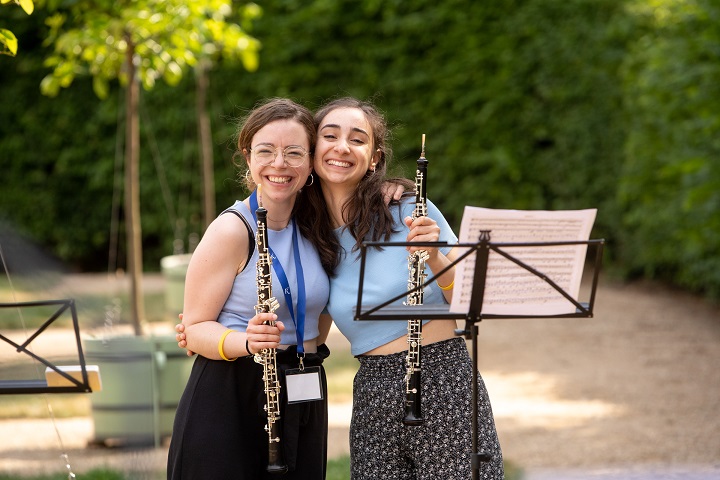 Zwei Klarinettistinnen mit ihren Instrumenten im Rheinsberger Schlosspark. Beide lachen und halten ihre Instrumente in die Kamera.